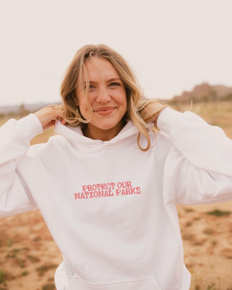 May include: A white long-sleeve hooded sweatshirt featuring the text "PROTECT OUR NATIONAL PARKS" printed in a distressed red font across the chest. The garment includes a hood with drawstrings and a front kangaroo pocket for storage.