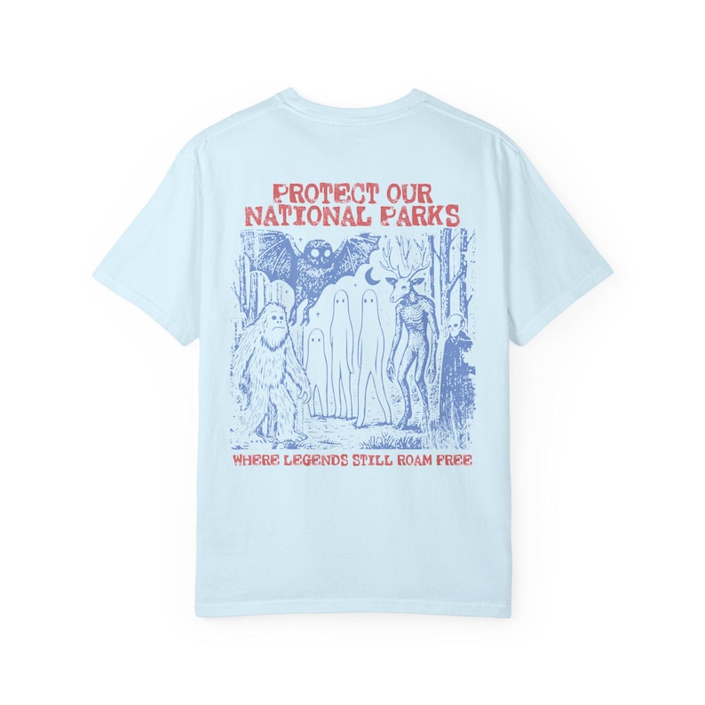 May include: A light blue t-shirt featuring a blue graphic print on the back. The design depicts a forest scene with a bat, a sasquatch, ghosts, a deer-like creature, and text that reads PROTECT OUR NATIONAL PARKS WHERE LEGENDS STILL ROAM FREE.