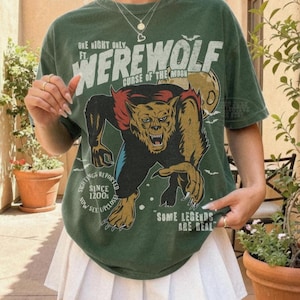 May include: A forest green short-sleeve t-shirt featuring a graphic of a werewolf with the text "ONE NIGHT ONLY FT. WEREWOLF CURSE OF THE MOON SIGHTINGS REPORTED SINCE 1200S NOW SEE UPCLOSE SOME LEGENDS ARE REAL" in white and yellow lettering.