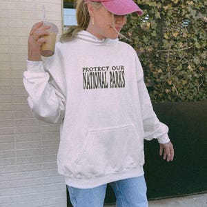 May include: A cream-colored hooded sweatshirt featuring the dark green text PROTECT OUR NATIONAL PARKS printed on the chest. The garment includes a front kangaroo pocket, long sleeves with ribbed cuffs, and a matching ribbed hemline.