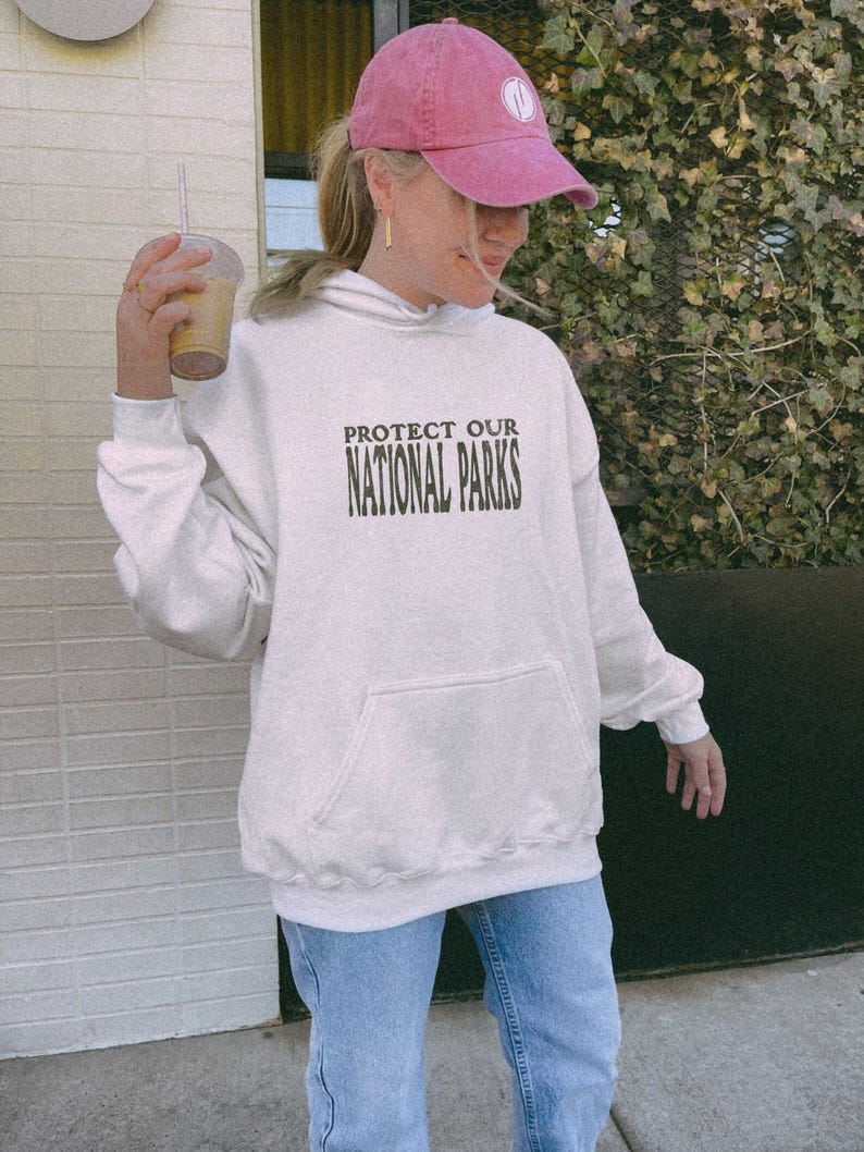 May include: A cream-colored hooded sweatshirt featuring the dark green text PROTECT OUR NATIONAL PARKS printed on the chest. The garment includes a front kangaroo pocket, long sleeves with ribbed cuffs, and a matching ribbed hemline.