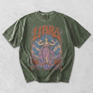 May include: An olive green short-sleeve t-shirt featuring a graphic print of a person in a purple dress holding scales. The text LIBRA is arched above the figure, with a blue starry background, orange floral accents, and a small Libra symbol below.