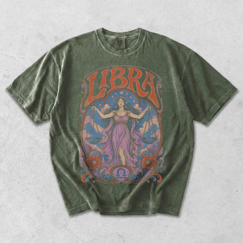 May include: An olive green short-sleeve t-shirt featuring a graphic print of a person in a purple dress holding scales. The text LIBRA is arched above the figure, with a blue starry background, orange floral accents, and a small Libra symbol below.