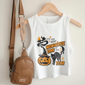 May include: White sleeveless crop top with a Halloween-themed graphic. The design features a jack-o'-lantern wearing a witch hat, a black cat, and the text "It's the most wonderful time of the year". A brown crossbody bag hangs to the left.