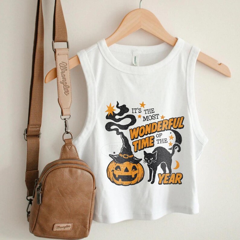 May include: White sleeveless crop top with a Halloween-themed graphic. The design features a jack-o'-lantern wearing a witch hat, a black cat, and the text "It's the most wonderful time of the year". A brown crossbody bag hangs to the left.