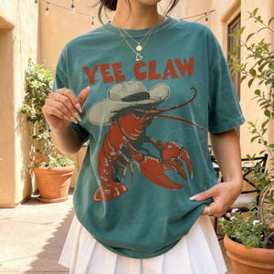 Op de afbeelding: Tealkleurig T-shirt met de tekst "YEE CLAW" in rode letters. De afbeelding is een rode kreeft met een cowboyhoed. De hoed is wit. Het T-shirt wordt gecombineerd met een witte geplooide rok.