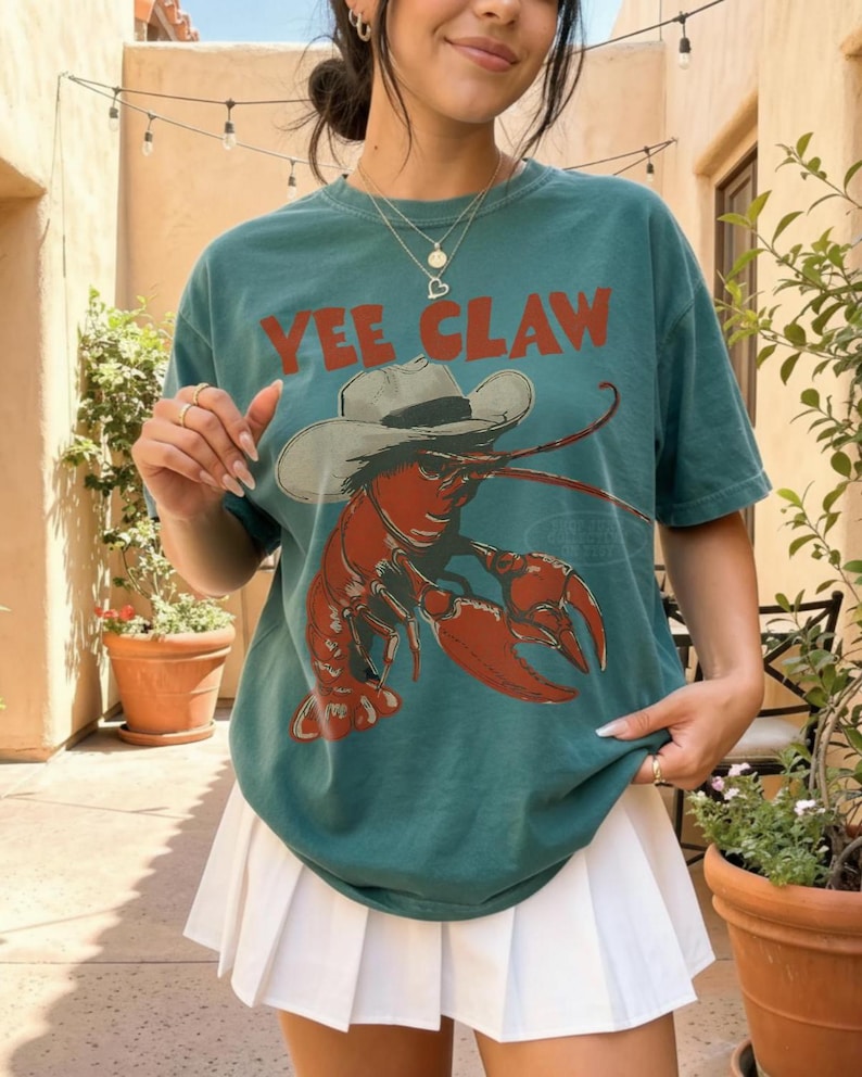 Op de afbeelding: Tealkleurig T-shirt met de tekst "YEE CLAW" in rode letters. De afbeelding is een rode kreeft met een cowboyhoed. De hoed is wit. Het T-shirt wordt gecombineerd met een witte geplooide rok.