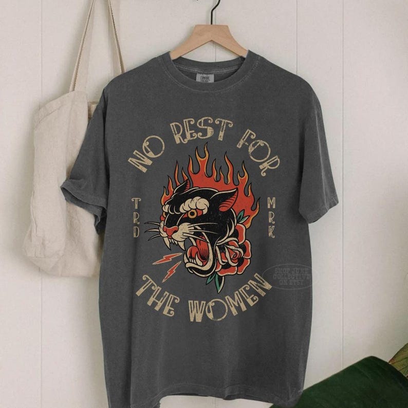 May include: Charcoal grey t-shirt with a retro graphic. The design includes a black panther with red flames, a rose, and the text "NO REST FOR THE WOMEN". The t-shirt is displayed on a wooden hanger.
