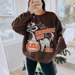 Peut inclure: Un sweat-shirt &agrave; col rond marron fonc&eacute; avec une impression graphique portant le texte IT'S THE MOST WONDERFUL TIME OF THE YEAR. Le motif comprend une tra&icirc;n&eacute;e de fum&eacute;e blanche, une citrouille d'Halloween avec un chapeau de sorci&egrave;re, un chat noir et des &eacute;toiles orange.