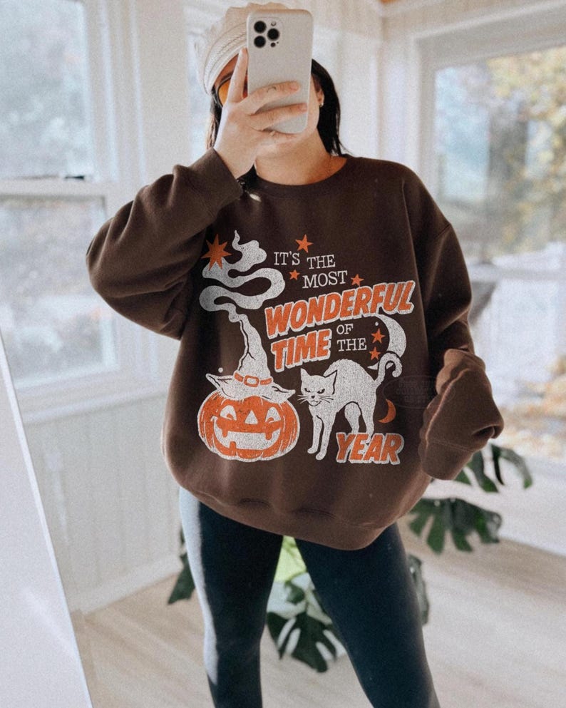 Peut inclure: Un sweat-shirt &agrave; col rond marron fonc&eacute; avec une impression graphique portant le texte IT'S THE MOST WONDERFUL TIME OF THE YEAR. Le motif comprend une tra&icirc;n&eacute;e de fum&eacute;e blanche, une citrouille d'Halloween avec un chapeau de sorci&egrave;re, un chat noir et des &eacute;toiles orange.