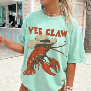 Op de afbeelding: Lichtgroen T-shirt met de tekst "YEE CLAW" in oranje. Het shirt heeft een cartoon illustratie van een rode kreeft met een cowboyhoed. Het T-shirt heeft een relaxte, oversized pasvorm.