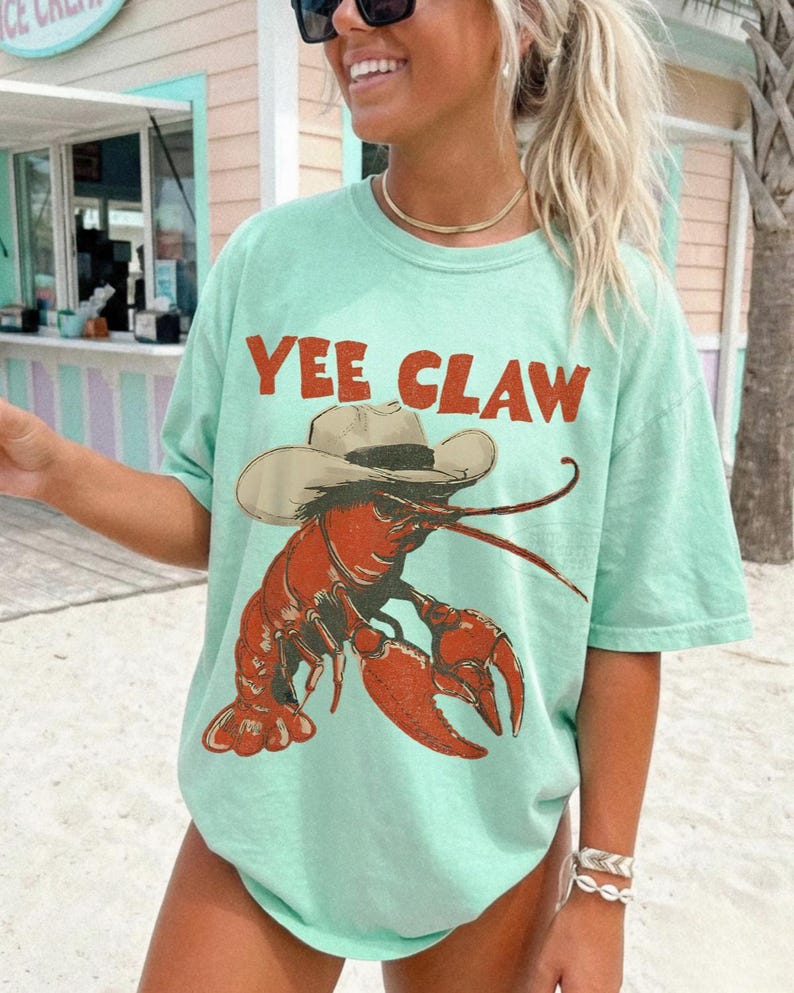 Op de afbeelding: Lichtgroen T-shirt met de tekst "YEE CLAW" in oranje. Het shirt heeft een cartoon illustratie van een rode kreeft met een cowboyhoed. Het T-shirt heeft een relaxte, oversized pasvorm.