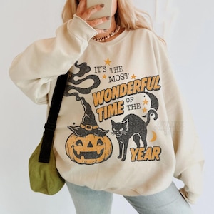Peut inclure: Un sweat-shirt beige &agrave; col rond avec une impression graphique portant le texte "IT'S THE MOST WONDERFUL TIME OF THE YEAR". Le motif comprend une citrouille d'Halloween avec un chapeau de sorci&egrave;re, un chat noir, des &eacute;toiles et un croissant de lune.
