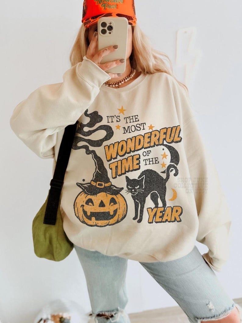 Peut inclure: Un sweat-shirt beige &agrave; col rond avec une impression graphique portant le texte "IT'S THE MOST WONDERFUL TIME OF THE YEAR". Le motif comprend une citrouille d'Halloween avec un chapeau de sorci&egrave;re, un chat noir, des &eacute;toiles et un croissant de lune.