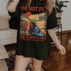 May include: A black t-shirt featuring a graphic of a bison in a woodland scene. The text "Do Not Pet The Fluffy Cows Yellowstone National Park" is printed on the shirt.