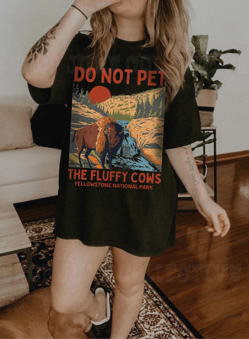 May include: A black t-shirt featuring a graphic of a bison in a woodland scene. The text "Do Not Pet The Fluffy Cows Yellowstone National Park" is printed on the shirt.