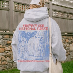 Op de afbeelding: Een lichtgrijze hoodie met een blauwe grafische print op de achterkant. De tekst luidt PROTECT OUR NATIONAL PARKS en WHERE LEGENDS STILL ROAM FREE. De illustratie toont een vleermuis, een sasquatch, geesten en een wezen met een hertenkop.