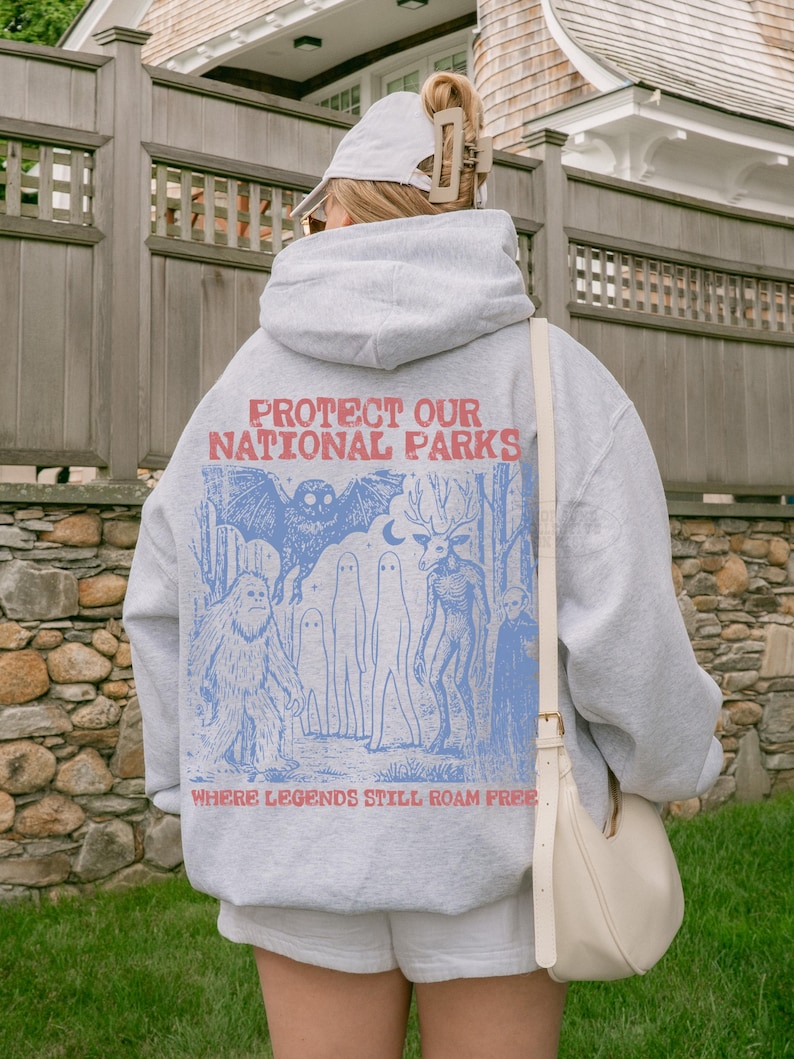 Op de afbeelding: Een lichtgrijze hoodie met een blauwe grafische print op de achterkant. De tekst luidt PROTECT OUR NATIONAL PARKS en WHERE LEGENDS STILL ROAM FREE. De illustratie toont een vleermuis, een sasquatch, geesten en een wezen met een hertenkop.