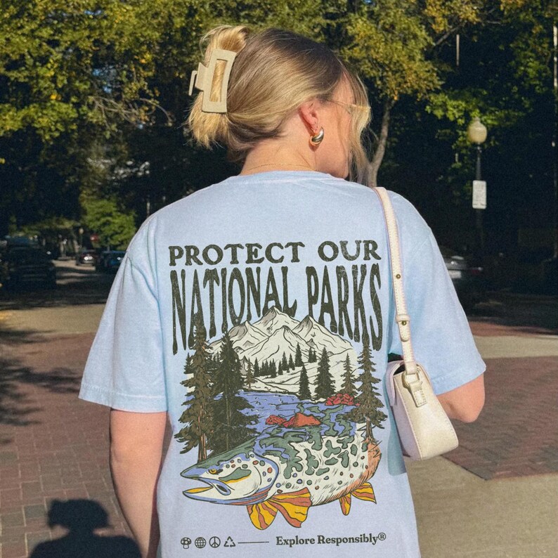 May include: Light blue t-shirt with a graphic print of a fish swimming in a lake with mountains and trees in the background. The text "Protect Our National Parks" is printed in black on the shirt. The text "Explore Responsibly" is printed in black at the bottom of the shirt.