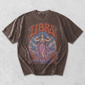May include: A dark brown short-sleeve t-shirt featuring a graphic print of a figure holding scales with the text LIBRA at the top. The design includes a central figure in a purple dress surrounded by blue clouds, stars, and orange floral accents.