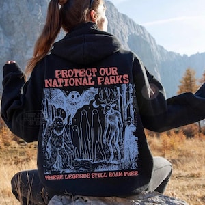 May include: A black hooded sweatshirt featuring a graphic print on the back with the text "PROTECT OUR NATIONAL PARKS" and "WHERE LEGENDS STILL ROAM FREE." The illustration depicts a forest scene with a large owl, a sasquatch, ghosts, and a deer.