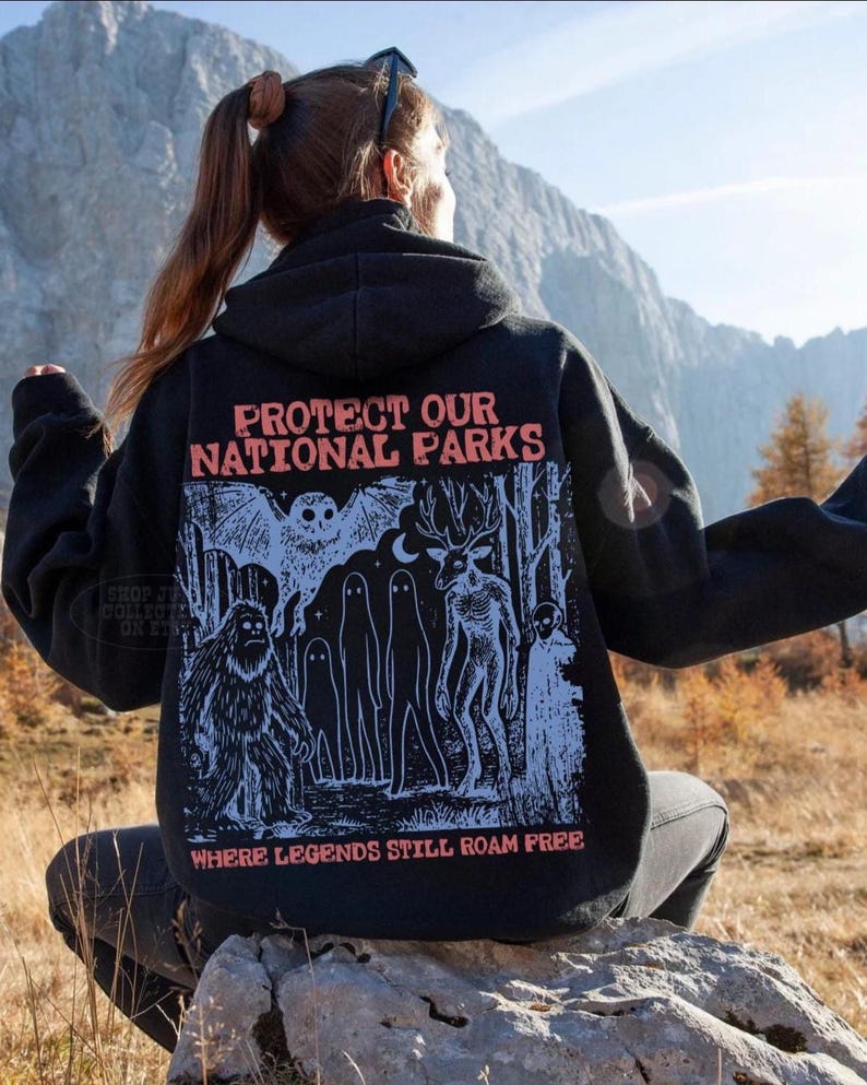 May include: A black hooded sweatshirt featuring a graphic print on the back with the text "PROTECT OUR NATIONAL PARKS" and "WHERE LEGENDS STILL ROAM FREE." The illustration depicts a forest scene with a large owl, a sasquatch, ghosts, and a deer.