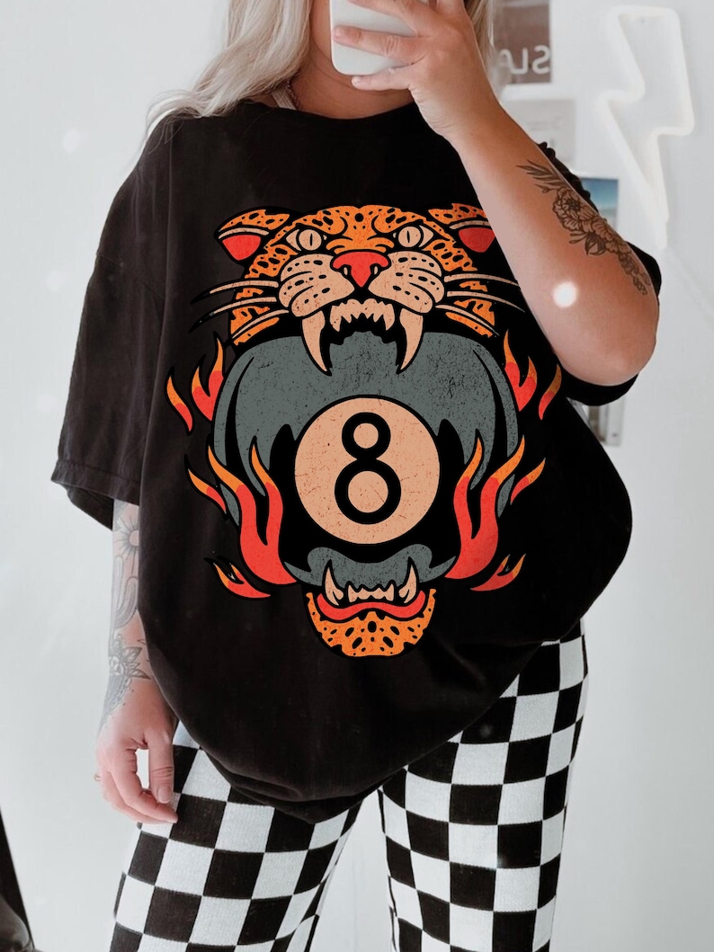 May include: Black t-shirt featuring a graphic of a snarling leopard biting an eight ball, surrounded by flames. The design is in shades of orange, red, and gray. The person is wearing black and white checkered pants.
