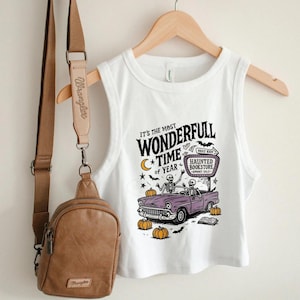 May include: White crop top with a Halloween-themed graphic. The design features a purple car with skeletons, pumpkins, and the text "It's the Most Wonderfull Time of Year." A brown crossbody bag hangs on the left.