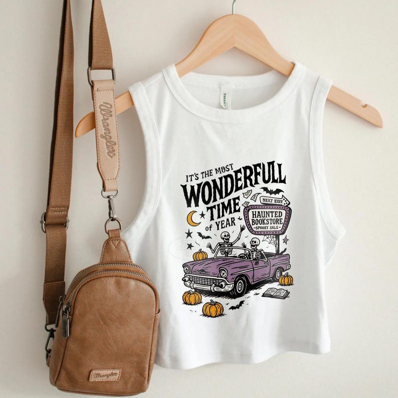 May include: White crop top with a Halloween-themed graphic. The design features a purple car with skeletons, pumpkins, and the text "It's the Most Wonderfull Time of Year." A brown crossbody bag hangs on the left.