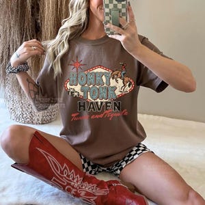 Pode incluir: Camiseta marrom com um gr&aacute;fico "Honky Tonk Haven" com um cowboy a cavalo, uma estrela e as palavras "Tune and Tequila". A pessoa est&aacute; usando botas de cowboy vermelhas e um bon&eacute; de beisebol marrom e branco.