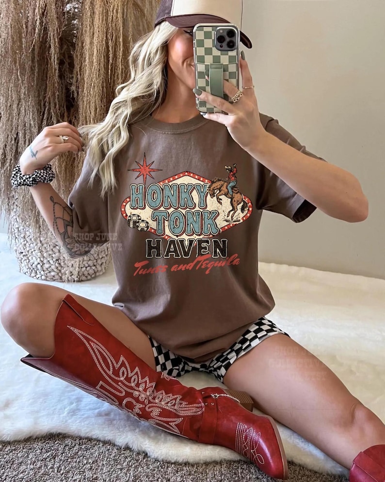 Pode incluir: Camiseta marrom com um gr&aacute;fico "Honky Tonk Haven" com um cowboy a cavalo, uma estrela e as palavras "Tune and Tequila". A pessoa est&aacute; usando botas de cowboy vermelhas e um bon&eacute; de beisebol marrom e branco.