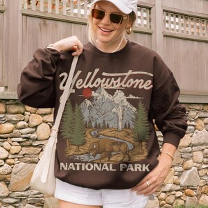 May include: A dark brown crewneck sweatshirt featuring a graphic print of Yellowstone National Park. The design includes a bison, pine trees, mountains, a geyser, and a red sun, with the text Yellowstone National Park printed in a vintage-style font.