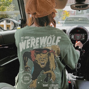 May include: An olive green t-shirt featuring a graphic print of a werewolf on the back. The text reads: One Night Only Ft. Werewolf Curse of the Moon, Sightings Reported Since 1200s, Now See Upclose, Some Legends Are Real. A brown corduroy cap is worn.
