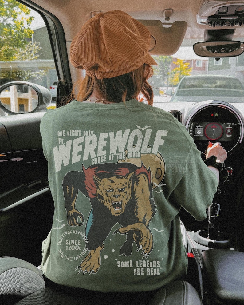 May include: An olive green t-shirt featuring a graphic print of a werewolf on the back. The text reads: One Night Only Ft. Werewolf Curse of the Moon, Sightings Reported Since 1200s, Now See Upclose, Some Legends Are Real. A brown corduroy cap is worn.