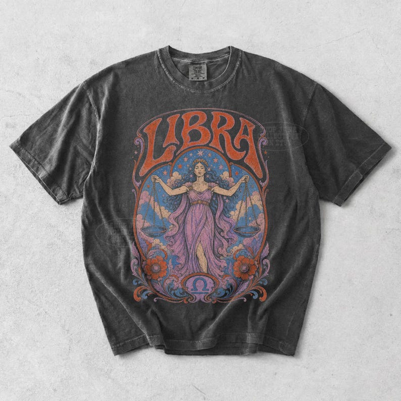 May include: A charcoal gray short-sleeve t-shirt featuring a graphic print of the Libra zodiac sign. The design displays a central figure holding scales, surrounded by clouds, stars, and floral motifs in shades of orange, purple, and blue.
