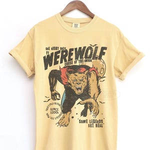 May include: A pale yellow short-sleeve t-shirt featuring a graphic of a werewolf with red accents and a full moon. The text reads: ONE NIGHT ONLY FT. WEREWOLF CURSE OF THE MOON, SIGHTINGS REPORTED SINCE 1200S NOW SEE UPCLOSE, and SOME LEGENDS ARE REAL.