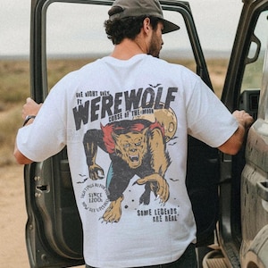 May include: A white short-sleeve t-shirt featuring a graphic on the back of a werewolf with red hair, a yellow moon, and bats. The text reads: ONE NIGHT ONLY FT. WEREWOLF CURSE OF THE MOON, SIGHTINGS REPORTED SINCE 1200s NOW SEE UPCLOSE, SOME LEGENDS ARE REAL.