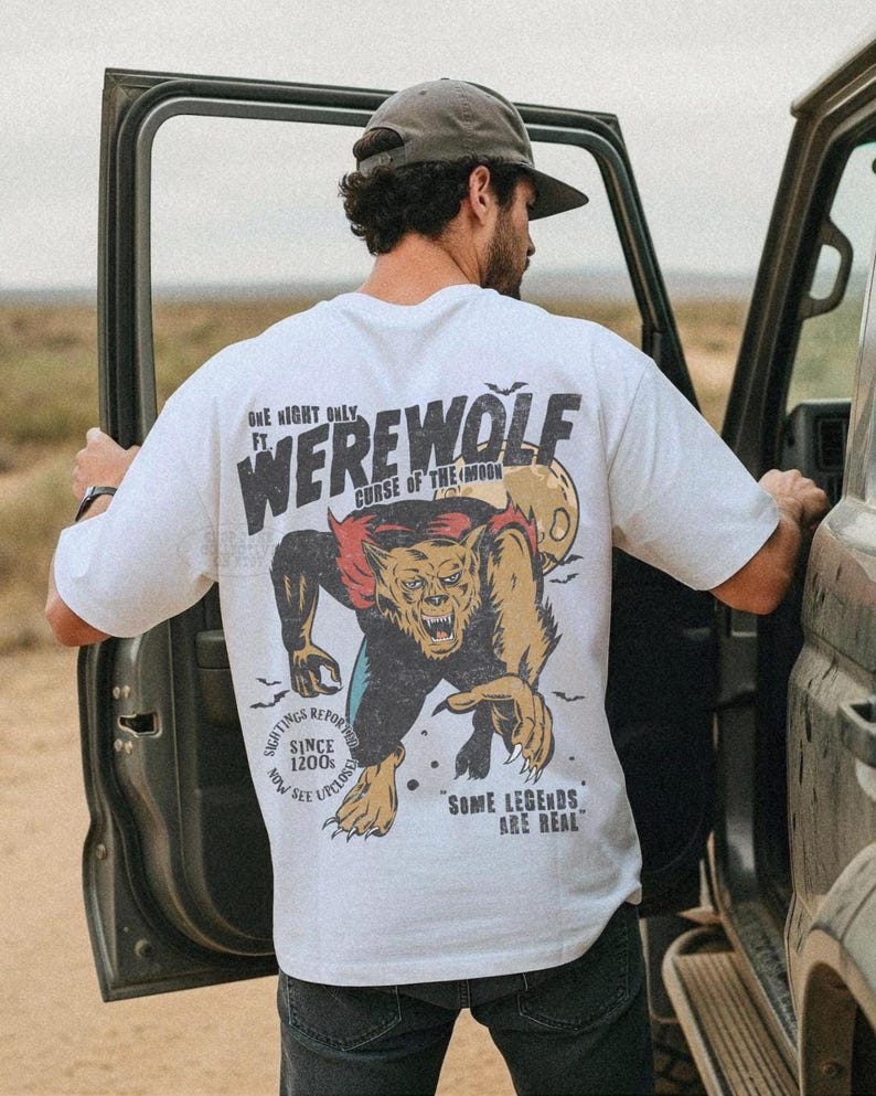 May include: A white short-sleeve t-shirt featuring a graphic on the back of a werewolf with red hair, a yellow moon, and bats. The text reads: ONE NIGHT ONLY FT. WEREWOLF CURSE OF THE MOON, SIGHTINGS REPORTED SINCE 1200s NOW SEE UPCLOSE, SOME LEGENDS ARE REAL.