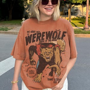 May include: A rust-colored short-sleeve t-shirt featuring a graphic print of a werewolf with the text "ONE NIGHT ONLY FT. WEREWOLF CURSE OF THE MOON" and "SIGHTINGS REPORTED SINCE 1200S NOW SEE UPCLOSE" along with "SOME LEGENDS ARE REAL".