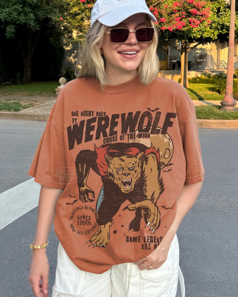 May include: A rust-colored short-sleeve t-shirt featuring a graphic print of a werewolf with the text "ONE NIGHT ONLY FT. WEREWOLF CURSE OF THE MOON" and "SIGHTINGS REPORTED SINCE 1200S NOW SEE UPCLOSE" along with "SOME LEGENDS ARE REAL".