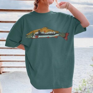 Puede incluir: Camiseta de color verde azulado con una ilustraci&oacute;n detallada de un pez. El dise&ntilde;o del pez incluye una escena de monta&ntilde;a y puesta de sol en amarillo, naranja, verde y rojo. La camiseta tiene un corte holgado.