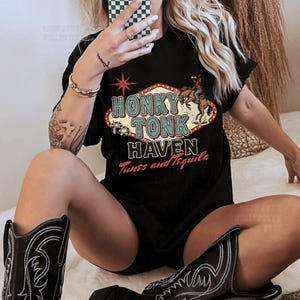 Pode incluir: Camiseta preta com a frase "Honky Tonk Haven Tunes and Tequila" em um design retr&ocirc;. A imagem tamb&eacute;m mostra botas de cowboy pretas com costuras brancas.