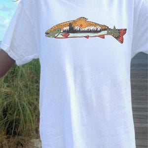 Puede incluir: Camiseta blanca con una ilustraci&oacute;n detallada de un pez. El pez tiene un cuerpo marr&oacute;n y naranja, una cola roja y una cabeza verde. El dise&ntilde;o del paisaje dentro del pez incluye monta&ntilde;as, &aacute;rboles y un sol poniente.