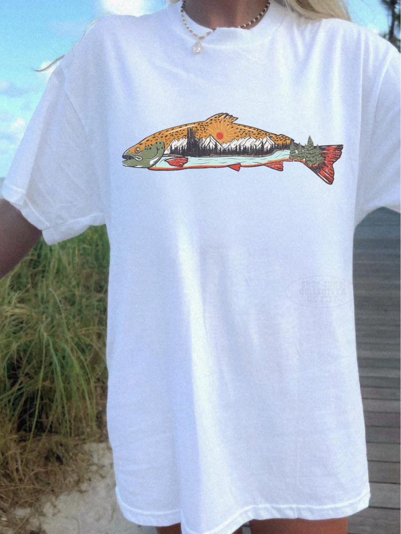 Puede incluir: Camiseta blanca con una ilustraci&oacute;n detallada de un pez. El pez tiene un cuerpo marr&oacute;n y naranja, una cola roja y una cabeza verde. El dise&ntilde;o del paisaje dentro del pez incluye monta&ntilde;as, &aacute;rboles y un sol poniente.