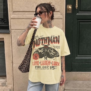 May include: A pale yellow short-sleeve t-shirt featuring a black moth graphic and red text that reads MOTHMAN, NO SISSIES ALLOWED, LIVE LAUGH LURK, FRI. DEC 15TH, POINT PLEASANT, WV 67, and a leopard print shoulder bag with a black and white strap.