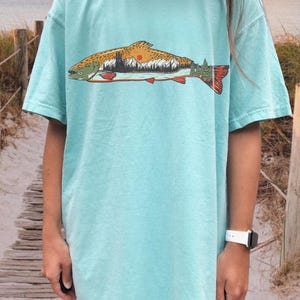 Puede incluir: Camiseta azul claro con un dise&ntilde;o de pez que contiene una escena de monta&ntilde;a y puesta de sol. El dise&ntilde;o del pez utiliza tonos marrones, naranjas y verdes. La camiseta tiene mangas cortas y un corte holgado. El dise&ntilde;o tiene un tema de naturaleza.