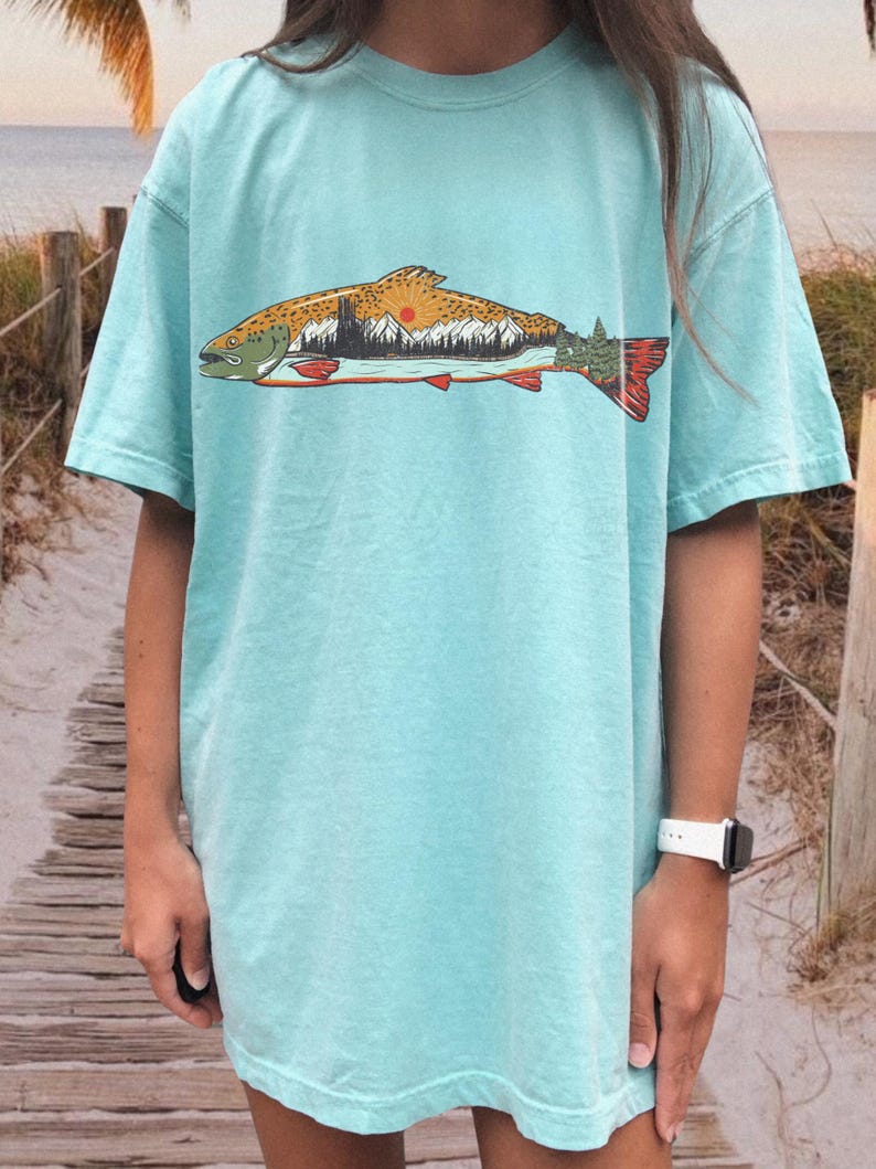 Puede incluir: Camiseta azul claro con un dise&ntilde;o de pez que contiene una escena de monta&ntilde;a y puesta de sol. El dise&ntilde;o del pez utiliza tonos marrones, naranjas y verdes. La camiseta tiene mangas cortas y un corte holgado. El dise&ntilde;o tiene un tema de naturaleza.