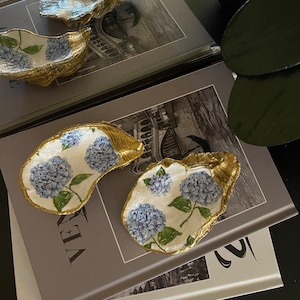 May include: Three gold oyster shell dishes with a white background and blue hydrangea floral pattern. The dishes are stacked on top of each other on a book.