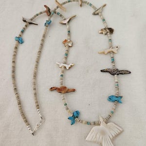 May include: A handcrafted necklace with animal and bird charms. The necklace has a silver clasp and is adorned with small, light-coloured beads, turquoise accents, and carved animal and bird pendants. The pendants include a bird, fish, and other animals.