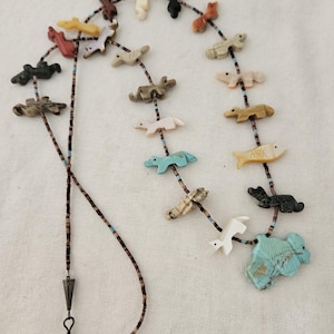 May include: A handcrafted necklace featuring animal-shaped beads in various colors, including turquoise, white, and brown. The beads are strung on a beaded cord with a metal clasp. The necklace showcases a unique, artistic design.